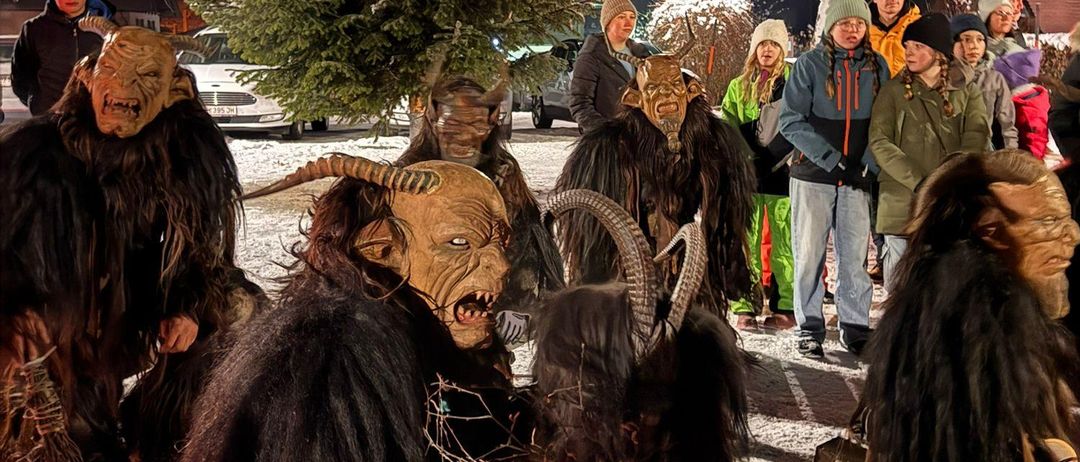 Eine Gruppe von Menschen in Hörnermasken steht um einen schneebedeckten Baum bei Nacht. Zuschauer in Winterkleidung beobachten von hinten. Ein Auto und eine Straßenlaterne sind im Hintergrund.