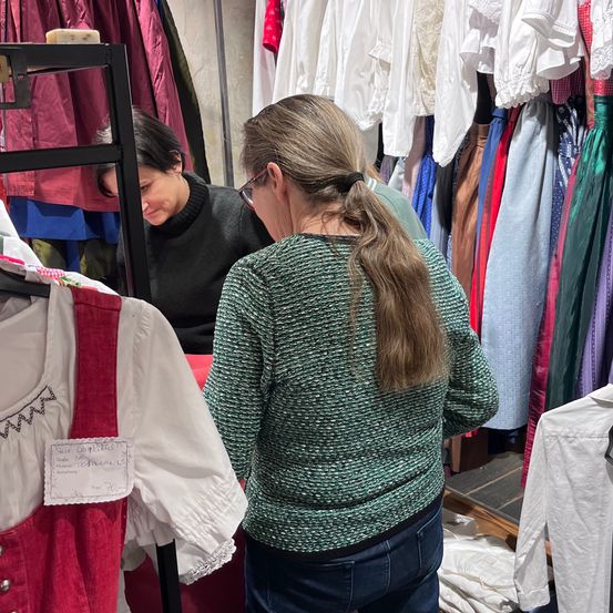 Eine Frau mit Brille schaut sich in einem Geschäft Kleidung an mit einer anderen Frau. Verschiedene Kleidungsstücke hängen an den Ständern.