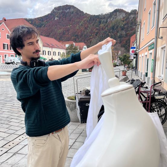 Ein Mann mit Kopfhörern justiert ein Kleidungsstück an einem Mannequin vor einer kleinen Stadt mit Bergen im Hintergrund.