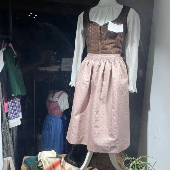 Ein traditionelles deutsches Kleid mit braunem Oberteil und rosa Rock, ausgestellt in einem Schaufenster, mit anderen traditionellen Kleidungsstücken im Hintergrund.