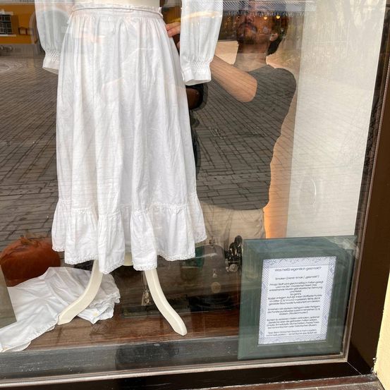 Ein weißes Kleid in einem Schaufenster ausgestellt, mit einem Schild, das fragt, ob Rauchen erlaubt ist. Ein Mann ist im Glas hinter dem Kleid reflektiert.