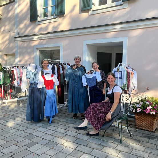 Fünf Frauen in traditioneller Kleidung stehen vor einem Gebäude und halten Kleider an Kleiderbügeln. Eine Frau sitzt auf einem Stuhl und lächelt. Vor ihnen hängen Kleider an Kleiderbügeln.