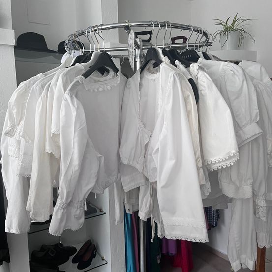 Ein Metallständer mit weißen Blusen hängt in einem Raum. Regale mit Schuhen und eine Topfpflanze sind sichtbar. Die Wand und Decke sind weiß.