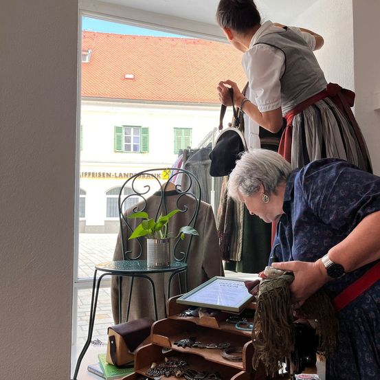 Zwei ältere Frauen untersuchen Gegenstände auf einem Tresen, einschließlich Schmuck, ein gerahmtes Zertifikat und eine Topfpflanze, mit Blick auf ein Gebäude mit rotem Dach und grünen Fensterläden durch das Fenster.