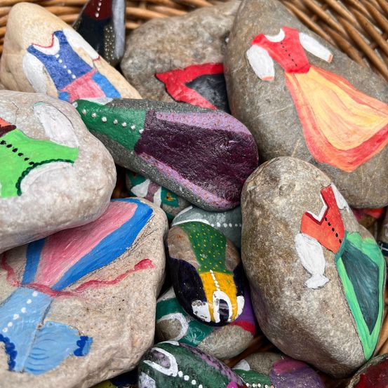 Ein Korb voller bunt bemalter Steine, die verschiedene Kostüme darstellen. Die Steine reichen von roten Kleidern bis hin zu grünen Westen und zeigen eine Vielzahl von Designs.
