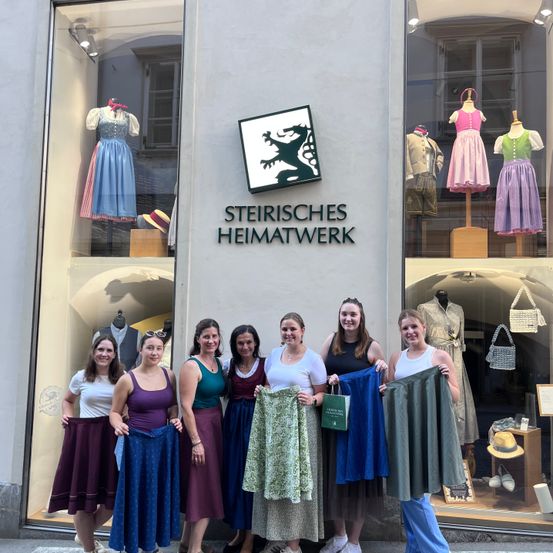 Eine Gruppe von Frauen steht vor einem Geschäft namens 'Steirisches Heimatwerk' und hält Röcke in verschiedenen Farben hoch. Mannequins und Kleidung sind in den Schaufenstern des Geschäfts ausgestellt.