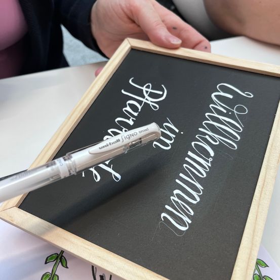 Eine Person schreibt mit einem weißen Stift auf eine schwarze Tafel. Die Tafel hat weiße Buchstaben, und die Person hält den Stift mit der linken Hand.