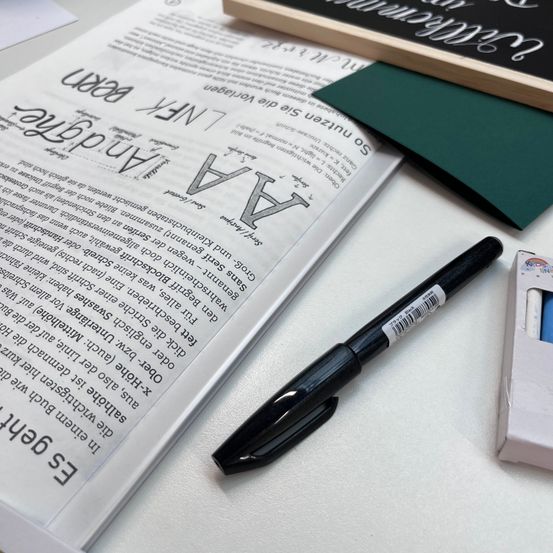 Ein Buch mit handgezeichneten Schriftproben, ein schwarzer Marker und ein schwarzes Tafel sind auf einem Schreibtisch.