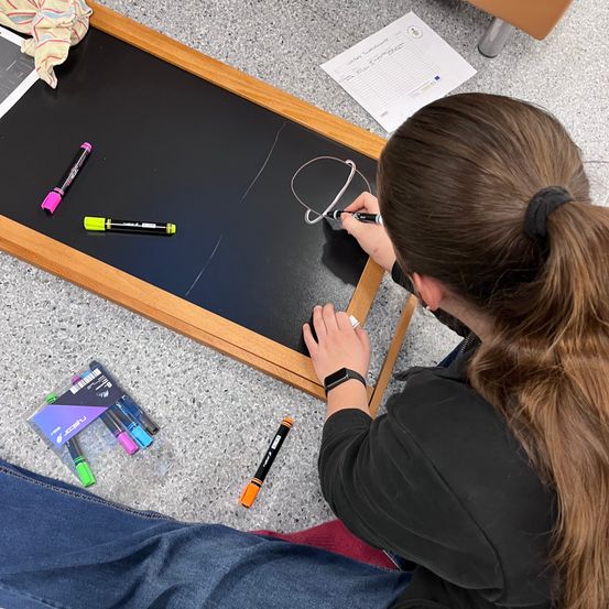Eine Frau mit einem Pferdeschwanz zeichnet mit Markern auf einer Tafel. Neben ihr liegen ein paar Marker und Papiere auf dem Boden.