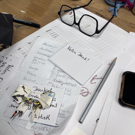 Ein Schreibtisch zeigt einen Papierfalter, eine Brille, einen Stift und ein Smartphone, mit handgeschriebenen Notizen und einer Karte, die 'Vielen Dank!' sagt.