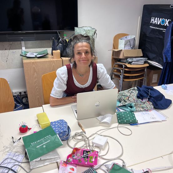 Eine Frau sitzt an einem Tisch mit einem Laptop und verschiedenen Büroartikeln. Sie lächelt und scheint zu arbeiten. Hinter ihr befinden sich Taschen, eine Schachtel und ein Fernseher an der Wand.