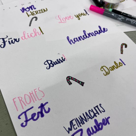 Ein weißes Papier mit verschiedenen handgeschriebenen Phrasen in verschiedenen Farben, einschließlich 'Love you!', 'Handmade', 'Bussi!', 'Frohes Fest', 'Danke!' und 'Weihnachtszauber'. Ein rosa und grüner Stift daneben.