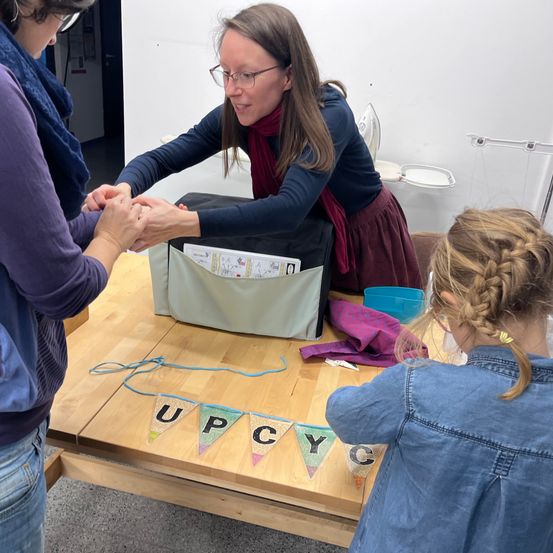 Drei Frauen in einer Werkstatt, eine hält die Hand der anderen, die beiden anderen beobachten. Ein Tisch mit einem Banner, das 'UPCYCLING' steht.