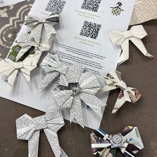 Auf einem Holztisch sind gefaltete Papierschlaufen aus Zeitungspapier zu sehen. Diese Schlaufen sind für Weihnachten gefertigt und zeigen verschiedene Designs und Farben.