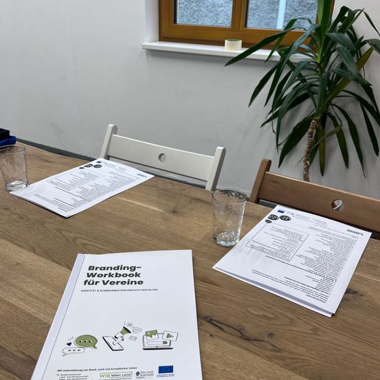 Ein hölzerner Tisch mit drei Papieren, zwei Gläsern und zwei Stühlen. Rechts vom Tisch steht eine Pflanze. Hinter dem Tisch befindet sich ein Fenster mit einem holzfarbenen Rahmen.