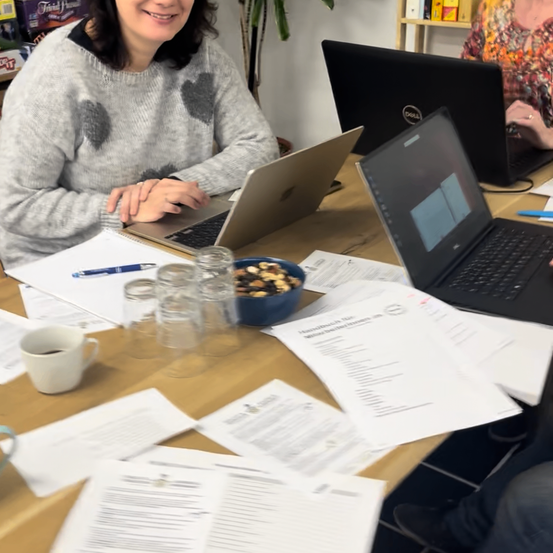Eine Frau lächelt, während sie an einem Tisch mit zwei Laptops sitzt, umgeben von Dokumenten, einer Schüssel mit Snacks und einer Tasse. Hinter ihr arbeitet eine andere Frau an einem Laptop.