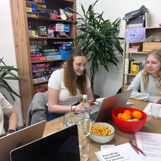 Drei Frauen sitzen um einen Tisch mit Laptops. Eine Frau lächelt und schaut auf ihren Laptop. Eine Schüssel mit Orangen und ein Glaskrug stehen auf dem Tisch. Bücher und Brettspiele im Hintergrund.