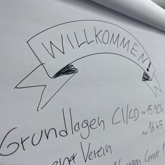 Eine weiße Tafel mit handgeschriebenem Text 'Willkommen!' und 'Grundlagen'. Unten steht ein Zeitplan für 'Verein' mit Zeiten 15:50 und 16:45. Oben rechts befindet sich ein Symbol.