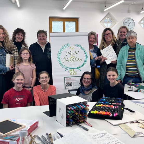 Eine Gruppe von Frauen und Mädchen versammelt sich um einen Tisch mit Kunstmaterialien, lächelt und posiert für ein Foto. Hinter ihnen befindet sich ein Banner mit den Worten 'Kraft und Tradition'.
