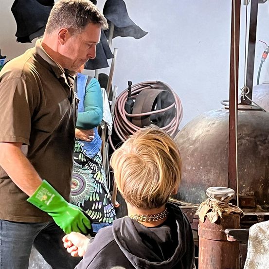 Ein Mann mit grüner Handschuh gibt einem kleinen Kind Anweisungen, was in einer Werkstatt mit Metallgeräten und Werkzeugen zu sehen scheint.