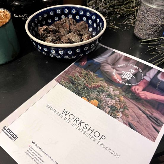 Ein Workshop-Prospekt liegt auf einem Tisch mit einer Schüssel mit getrockneten Kräutern, einem Glas Lavendel und einer Tasse. Der Prospekt steht für 'Workshop mit einheimischen Pflanzen'. Eine Person ist bei der Arbeit mit Pflanzen zu sehen.