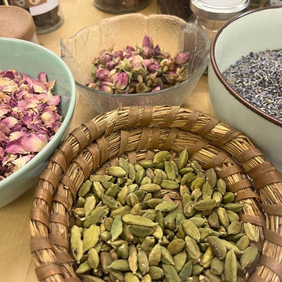 Verschiedene Kräuter und Gewürze in Schalen: lila Rosenblätter, rosa Blütenblätter, grüne Kardamomsamen und Lavendel. Ein Holztisch hält diese Gegenstände.