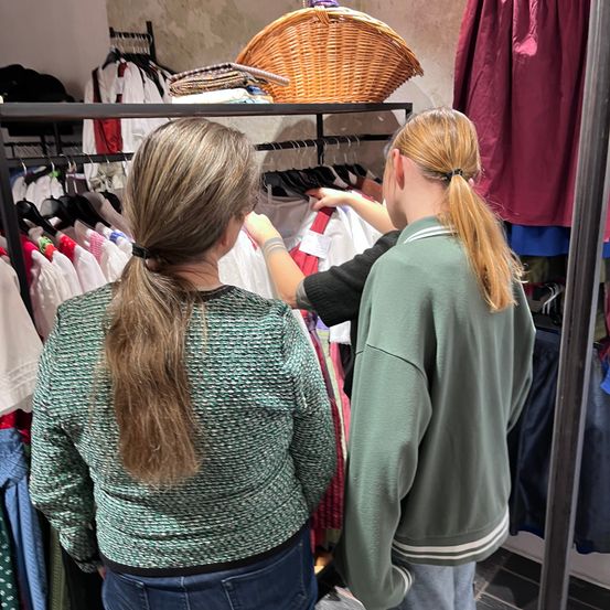 Zwei Frauen durchstöbern einen Bekleidungsladen. Eine Frau hat den Rücken zur Kamera gewandt, die andere hält einen Kleiderbügel. Kleidung hängt an Kleiderstangen, und ein Korb befindet sich auf einem Regal.