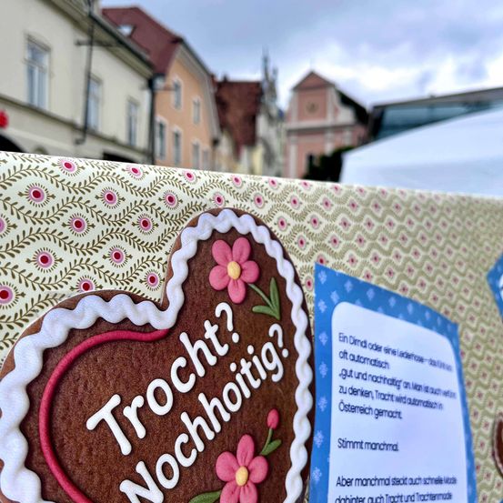 Ein herzförmiger Lebkuchen mit den Worten 'Trocken? Nochnoitig?' steht auf einer gemusterten Oberfläche. Dahinter sind Gebäude mit roten Dächern und ein bewölkter Himmel zu sehen. Eine blaue Karte befindet sich neben dem Lebkuchen.