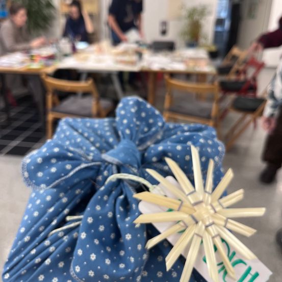 Ein blauer Beutel mit Blumenmuster und einem weißen Anhänger mit sternförmigem Design befindet sich in einem Raum mit Tischen und Stühlen. Die Tische haben Papiere und Gegenstände darauf. Es gibt verschwommene Personen, die an den Tischen sitzen.