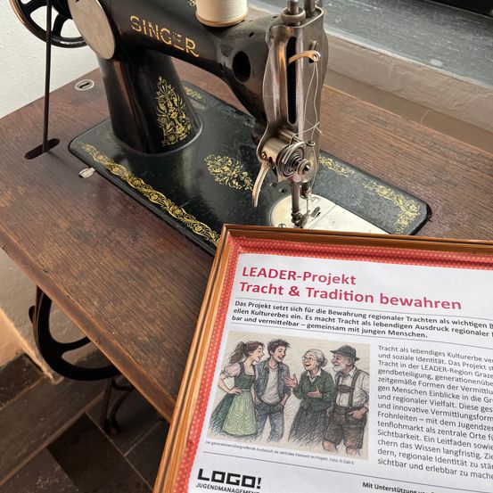 Eine alte Singer Nähmaschine auf einem Holztisch mit einem Flyer mit dem Titel 'LEADER-Projekt Tracht & Tradition bewahren' im Vordergrund. Der Flyer spricht über die Bewahrung regionaler Trachten als kulturelle Identität.
