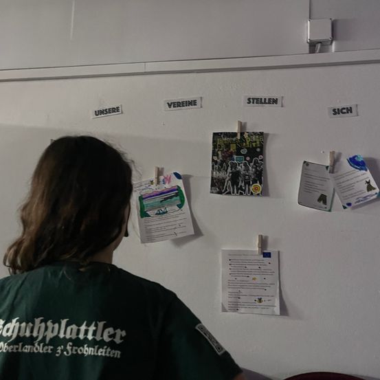 Eine Person in einem grünen Shirt schaut auf eine weiße Tafel mit mehreren angehefteten Papieren. Die Papiere haben Wörter wie 'unsere', 'vereine', 'stellen' und 'sich'. Die Papiere haben verschiedene Designs und Texte.