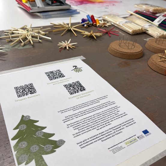 Ein Workshop-Setup mit Papier, bunten Strohhalmen und einem bedruckten Bogen mit Weihnachtsbaum-Design und QR-Codes, was auf eine Bastel- oder Bildungsaktivität hinweist.