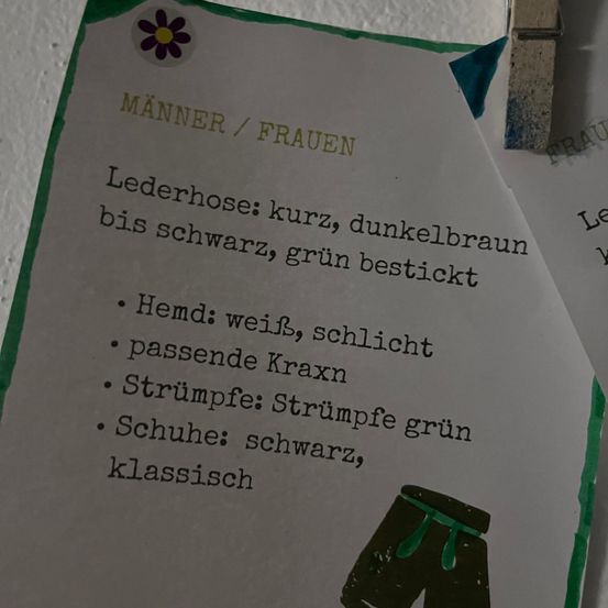 Ein an eine Wand geheftetes Papier beschreibt Männer- und Frauenkleidung. Für Männer: Shorts, Lederhose, weißes Hemd, Socken und schwarze Schuhe. Für Frauen: grüne Socken, grüne Shorts und grüne Socken.