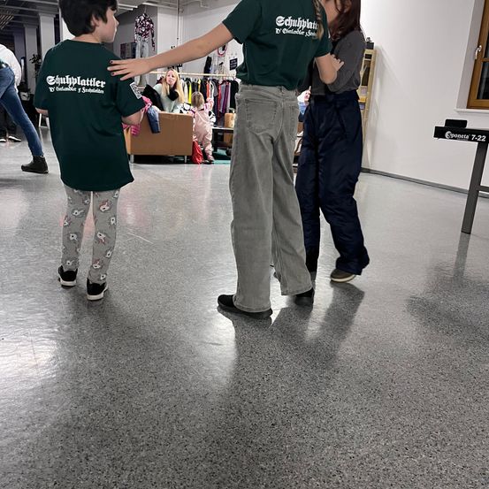 Ein Kind in einem grünen Shirt steht mit einer Frau, die seine Hand hält, in einem Raum mit grauem Boden. Dahinter unterhält sich eine Frau mit einem Kind auf einem Sofa.