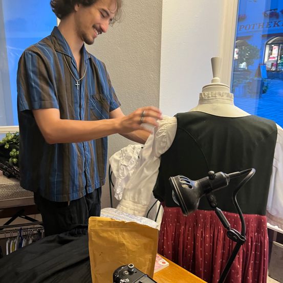 Ein Mann steht vor einem Mannequin und passt ein Kleidungsstück an. Er lächelt und trägt eine Halskette. Eine Lampe beleuchtet den Bereich, und Kleidung ist auf einem Tisch ausgestellt.