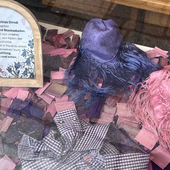 Ein violetter Hut mit breiter Krempe liegt auf einem Haufen bunter Stofffetzen. Ein hölzernes Schild im Hintergrund bietet Details zu Stoffresten.