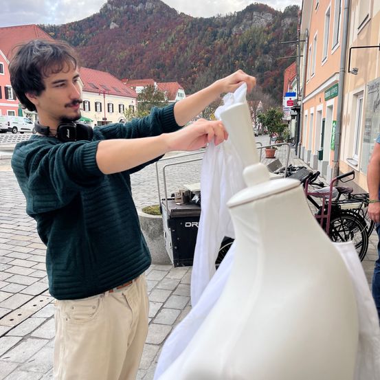 Ein Mann justiert ein weißes Kleidungsstück an einem Mannequin draußen auf einer Kopfsteinpflasterstraße. Hinter ihm sind Gebäude mit roten Dächern zu sehen, und ein Berg ist im Hintergrund.