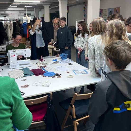 Eine Gruppe von Schülern lernt in einem Klassenzimmer zu nähen. Eine Frau unterrichtet sie, während sie vor einem Tisch mit verschiedenen Gegenständen steht.