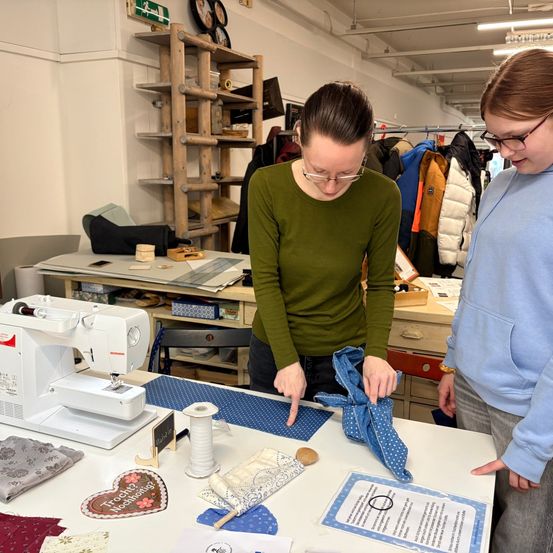 Eine Frau in einem grünen Shirt steht an einem Tisch in einem Raum mit einer Nähmaschine und Stoff. Eine andere Frau in einem blauen Shirt schaut sich ein Stück Stoff an.