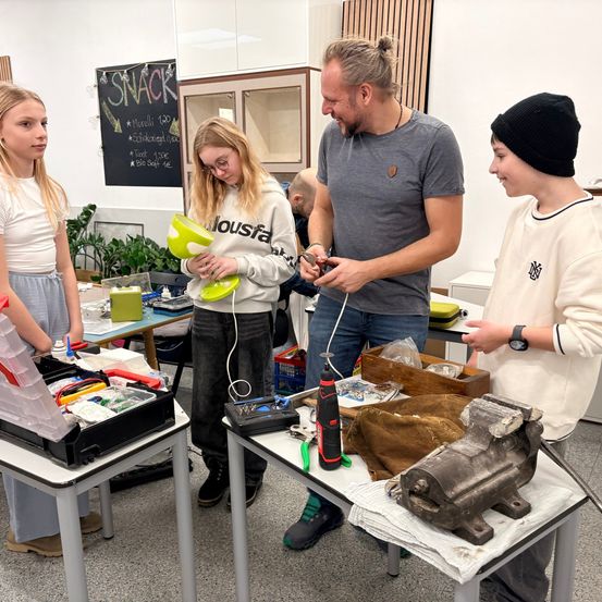 Eine Gruppe von Schülern arbeitet in einer Werkstatt an einem Projekt. Ein Mann erklärt ihnen etwas. Eine Frau hält eine grüne Schüssel und schaut den Mann an. Eine andere Frau schaut ebenfalls den Mann an.