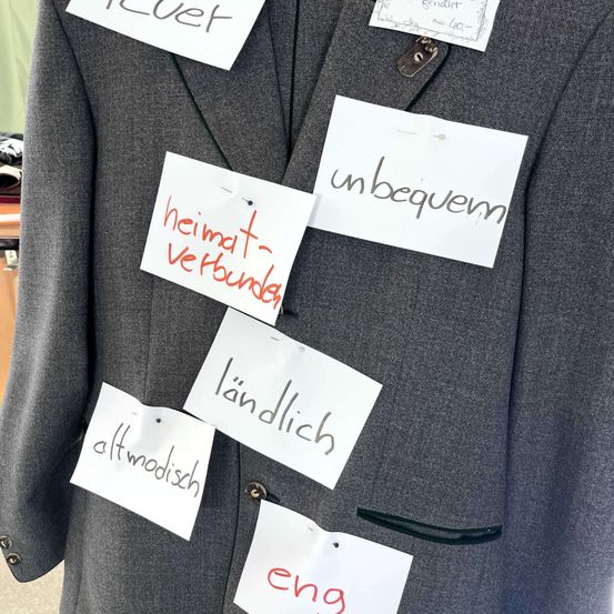 Ein grauer Mantel mit mehreren weißen Haftnotizen. Auf den Notizen stehen: 'unbequem,' 'heimat-verbunden,' 'ländlich,' 'altmodisch,' 'eng' und ein Etikett, das 'gewährt' mit der Nummer '40' liest.
