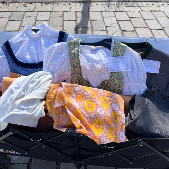 Eine Sammlung verschiedener Kleidungsstücke ist in einem schwarzen Wagen ausgestellt. Dazu gehören weiße Blusen, ein Blumenkleid und eine graue Jacke. Einige Artikel haben Preisschilder angebracht.