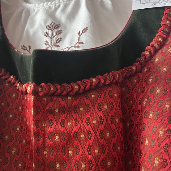 Ein Nahaufnahme eines roten und schwarzen gemusterten Kleides mit rotem Saum und grüner Futter. Im Hintergrund ist ein gestickter weißer Stoff mit roten Blumendesigns zu sehen.