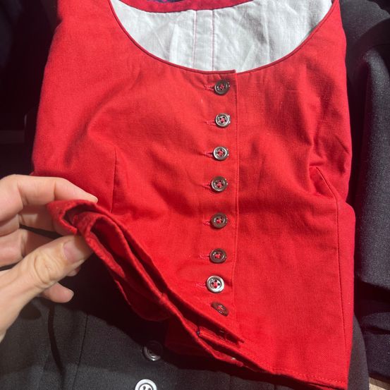 Ein Ausschnitt eines roten Kleidungsstücks mit weißem Innenfutter wird gezeigt. Es hat einen runden Ausschnitt und wird von einer Hand gehalten. Es hat insgesamt acht silberne Knöpfe.