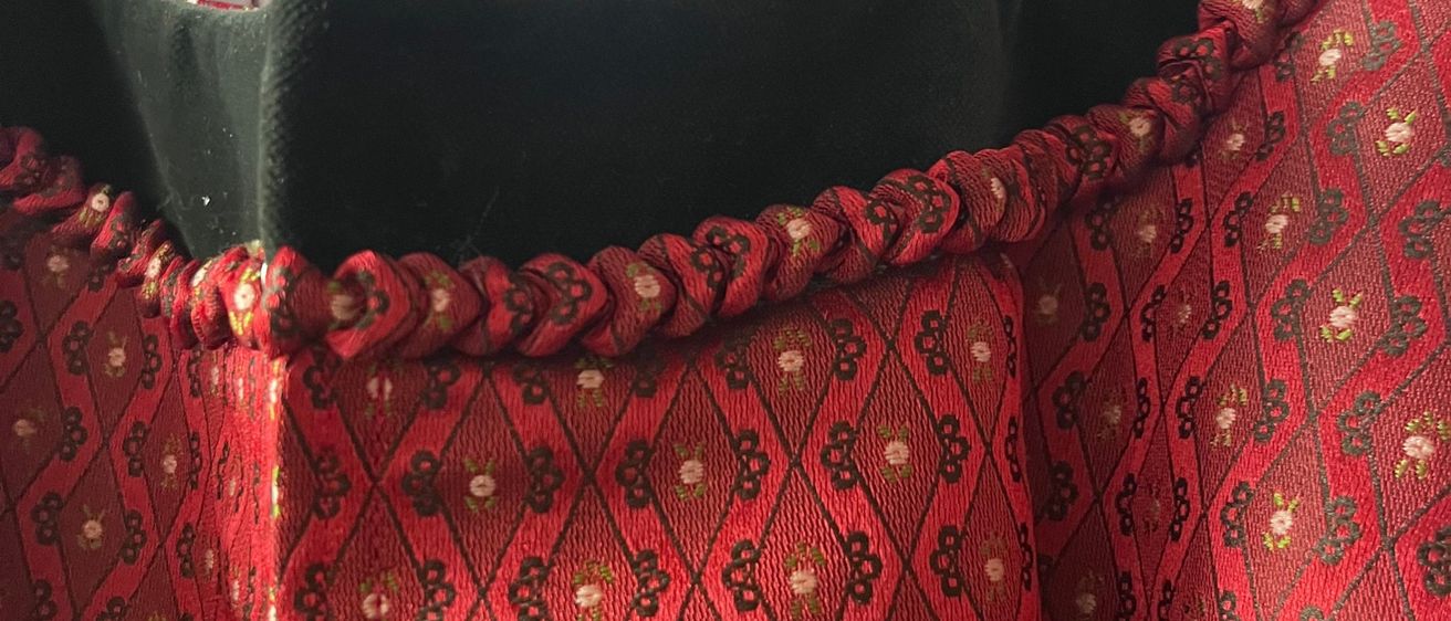 Ein Ausschnitt eines roten Samtkleides mit Blumenmustern und einem grünen Kragen. Das Kleid zeigt aufwendige Stickerei, und ein Teil eines weißen Kleidungsstücks ist im Hintergrund zu sehen.