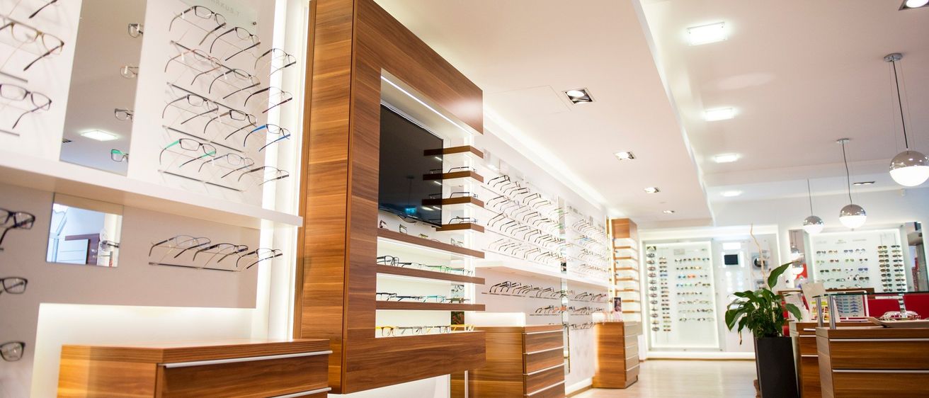 Der Innenraum eines Optikgeschäfts mit hölzernen Schränken mit mehreren Regalen, die verschiedene Brillen präsentieren. An den Wänden sind Brillen montiert, und auf einem der Schränke befindet sich ein Flachbildfernseher. Die Decke ist weiß mit Einbauleuchten.