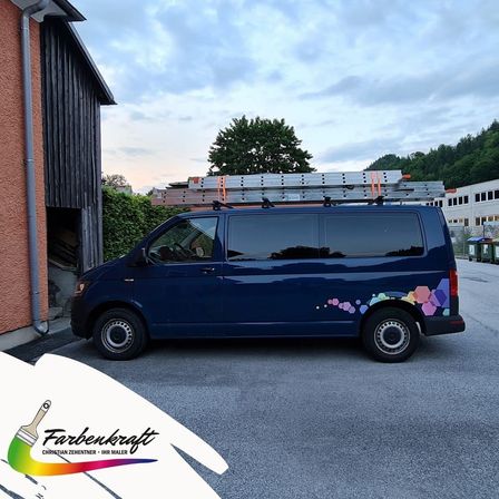 Ein blauer Van mit bunten sechseckigen Aufklebern steht in der Nähe eines Gebäudes. Eine Leiter ist auf dem Dach. Bäume und ein Gebäude sind im Hintergrund.