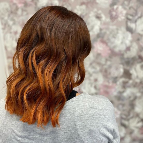 Eine Frau mit langen, welligen, rotbraunen Haaren in lockeren Wellen, die ein graues Shirt trägt und vor einem Blumenhintergrund steht.