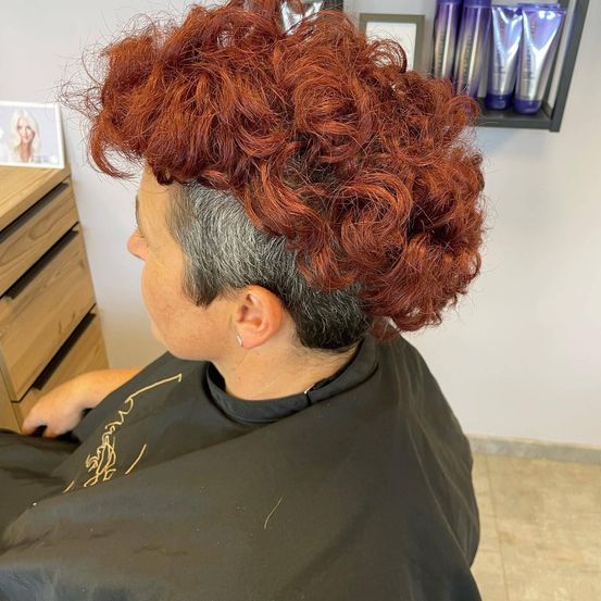 Eine Frau mit roten lockigen Haaren sitzt in einem Salon und trägt einen schwarzen Umhang. Hinter ihr sind ein Holzschrank und ein Regal mit Haarpflegeprodukten zu sehen.