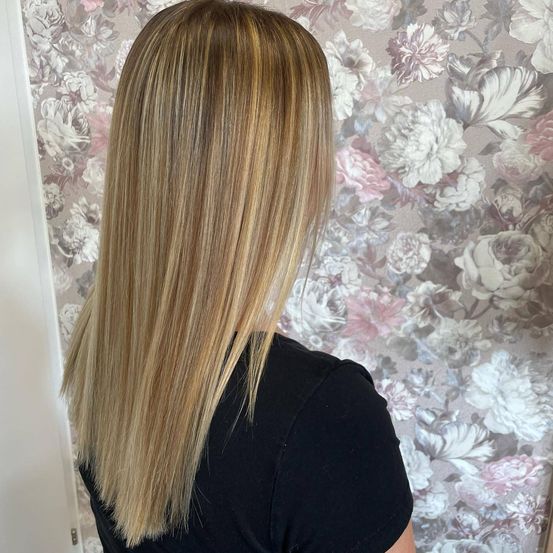 Eine Frau mit langen, blonden Haaren in lockeren Wellen steht vor einer floralen Tapete. Sie trägt ein schwarzes Oberteil.